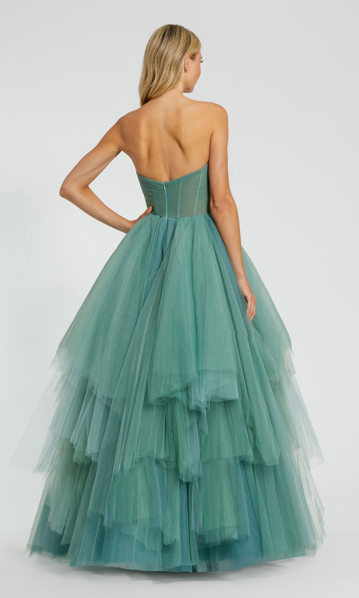 A woman stands with her back to the camera wearing the Long Formal Dress 20772 by Mac Duggal, a strapless gown with a ruched bodice and a full, tiered teal tulle skirt.